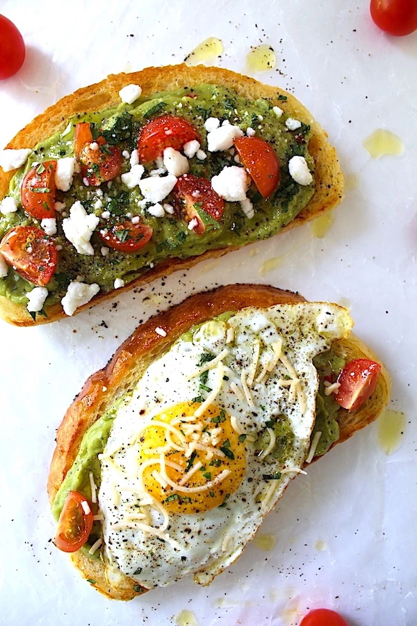 Two avocado toasts side by side, covered in fresh herbs, and drizzled olive oil.