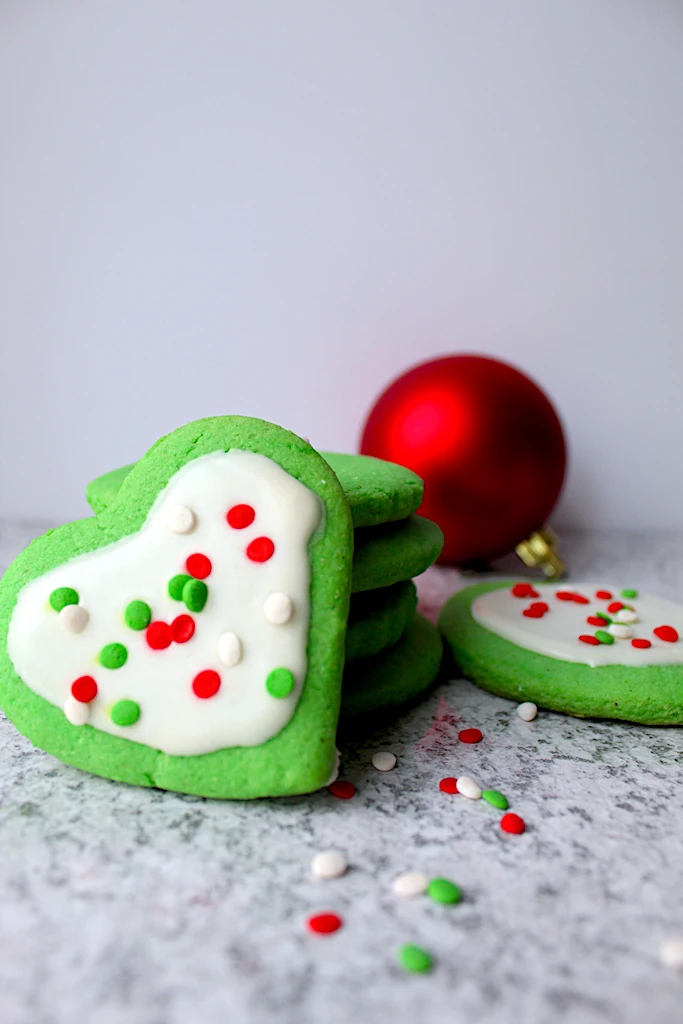 Green Grinch Inspired Almond Sugar Cookies with cute Christmas sprinkles and sugar cookie icing. A single red christmas ornament is seen in the back, reflecting a red glow onto the counter by the bright green cookies.